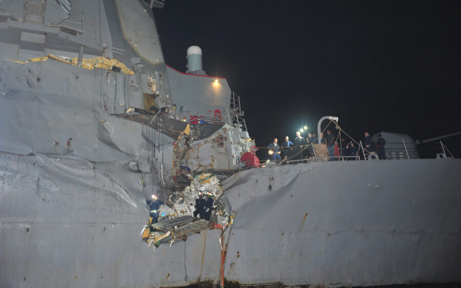 Een tankschip vaart door de Straat van Hormuz met militaire schepen op de achtergrond